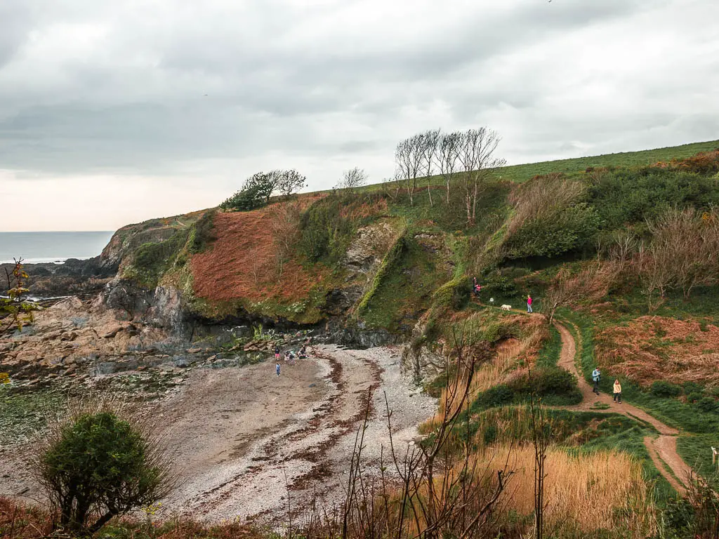 A beach cove surrounded by a grass covered cliff on the walk from Par to Fowey. There is a trail running across the grass cliff top with a few people walking on it.