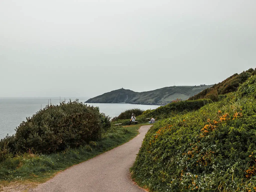 A path with a hedge to the left and a bush to the right and view out to sea ahead and the Rame Head Peninsular, on the walk route from Kingsand. There are a couple of people sitting down on the green ahead, next to the path. 