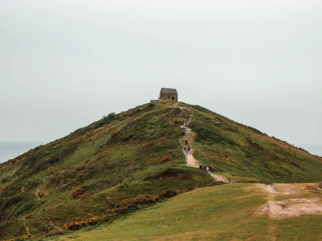 Looking to the big pointy hill of Rame Head with lots of people walking up it along the path. There is a small church at the top of the hill.