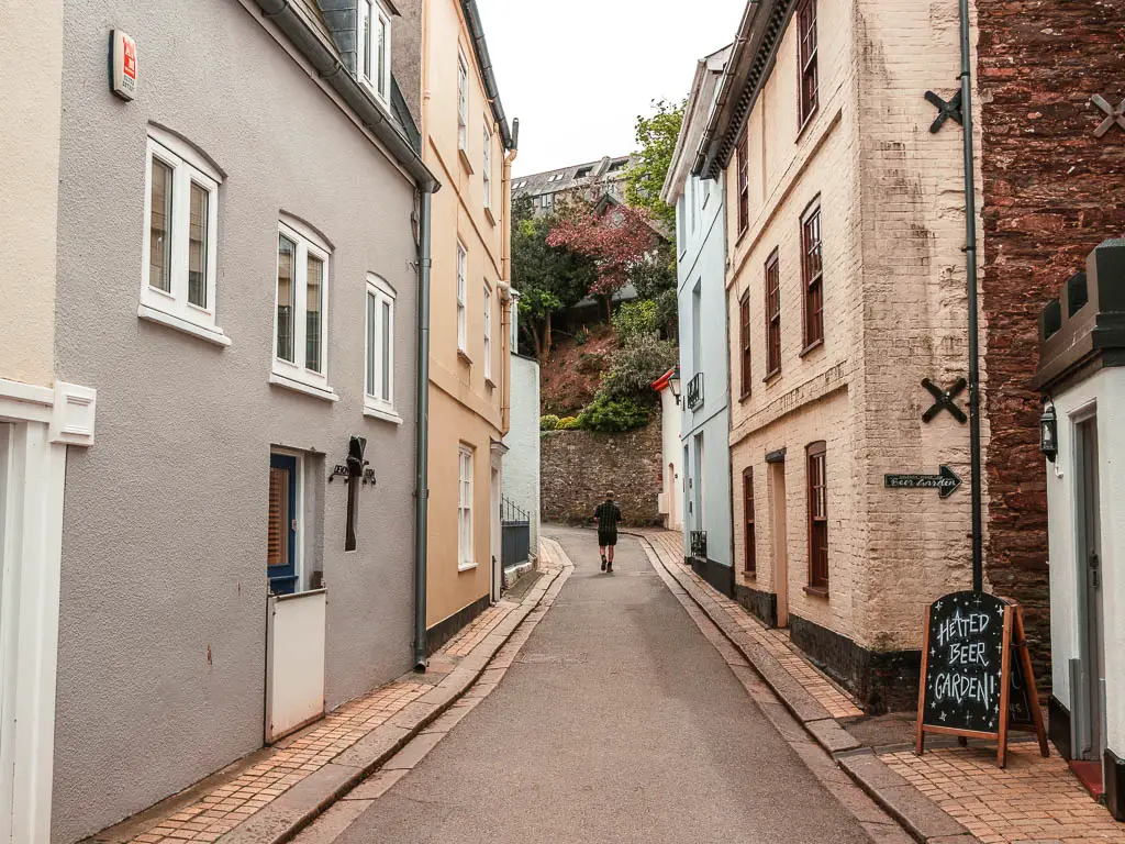 Looking along a narrow road between the pastel coloured houses at the start of the walk from Kingsand to Rame Head. There is a man walking ahead on the road. 