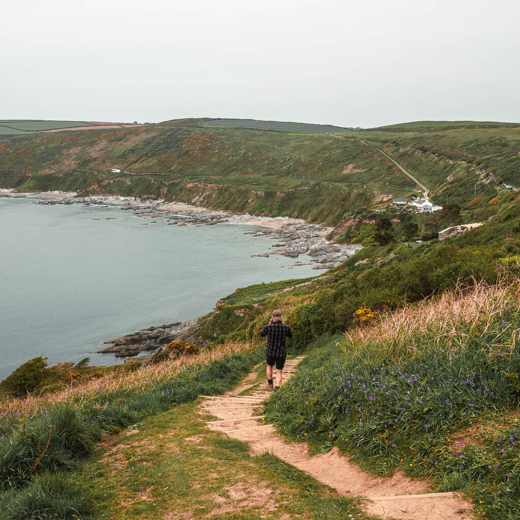 A man walking down the trail on the edge of the hill with a view to the sea and coastline ahead. 