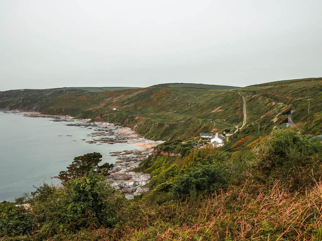 Looking across the grass covered cliff coastline on the Rame Head Peninsular walk. There are rocks in the sea at the bottom of the cliff, and the cliff leads up to a hilly clifftop. There is a small white house in one of the hill valleys ahead. 