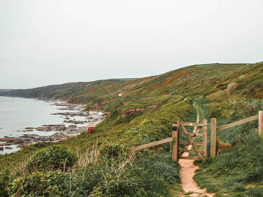 A narrow trail running straight through a wooden gate, with the gras covered cliff hillside ahead in the distance. 