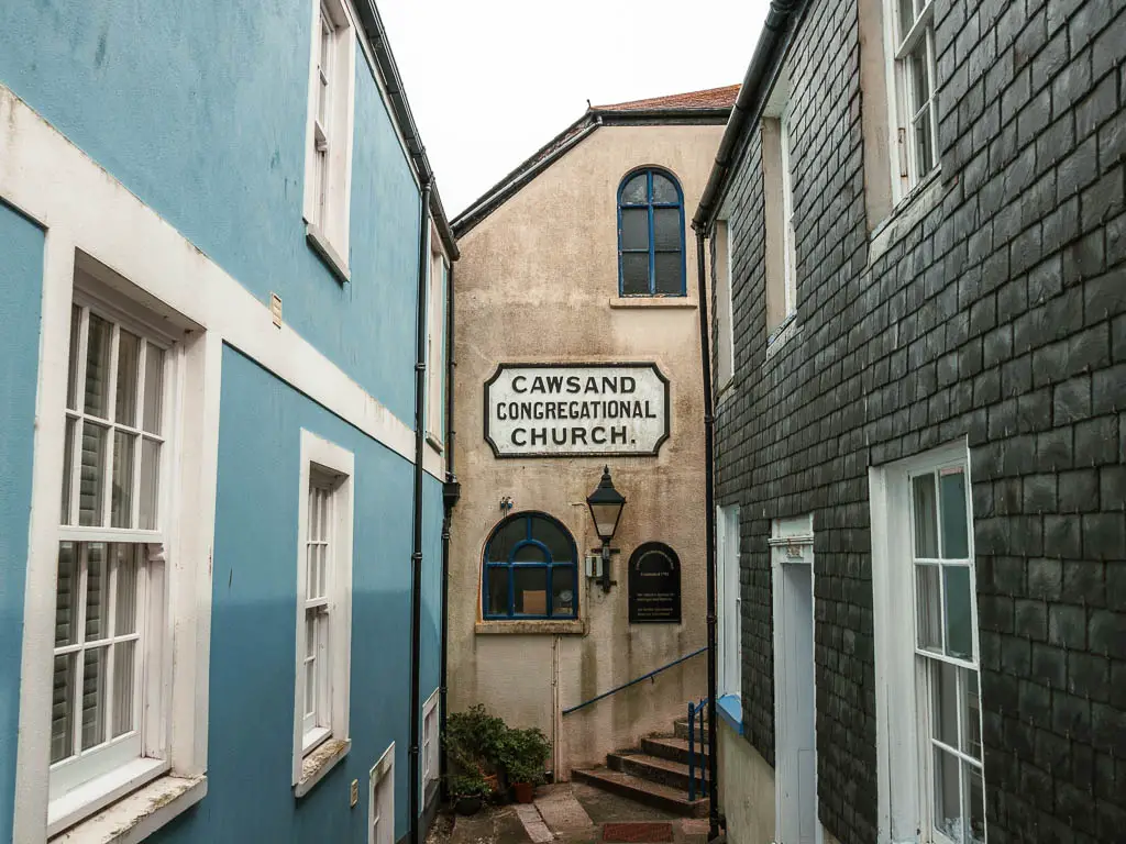 Looking down an alley between the houses in Caswsand. The left house is blue with white window frames, the right house is black with white window frames. Ahead is a cream coloured building with a church sign on the wall.