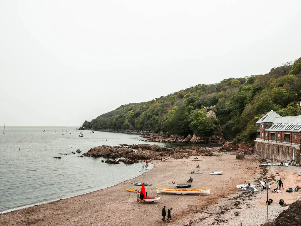 Looking down to the small beach of Cawsand, with the coastline ahead covered in trees. There are a few people on the beach. 