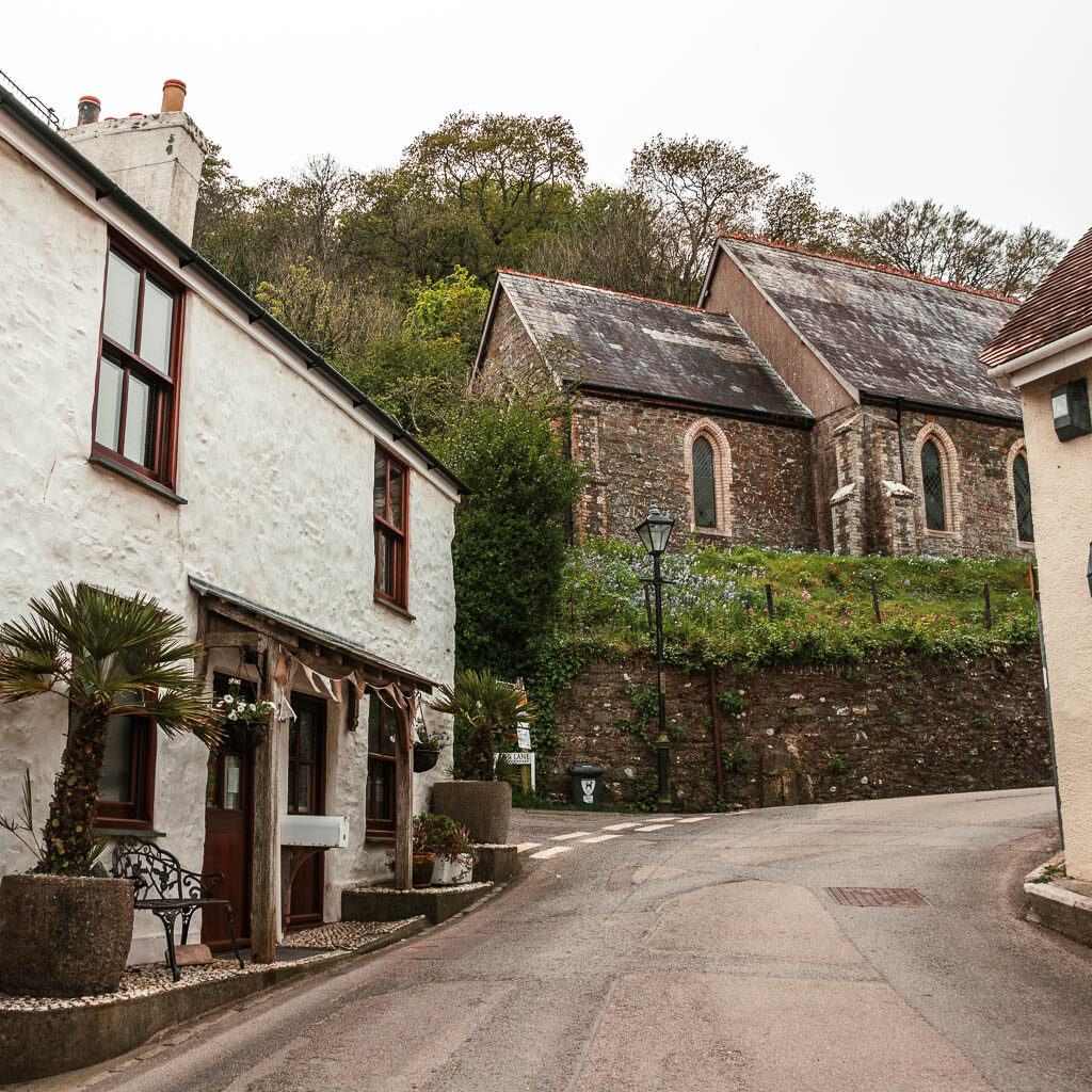 A road curving uphill to the right, with a white walled stone house on the left, and a church ahead. 