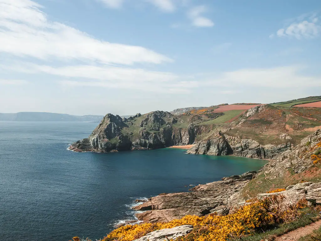 Looking down to the jagged rocky cliff cove from Prawle Point, on the walk from Salcombe. There are a few small beach coves visible, and the sea is deep blue turning to turquoise as it meets the beach and cliffs.