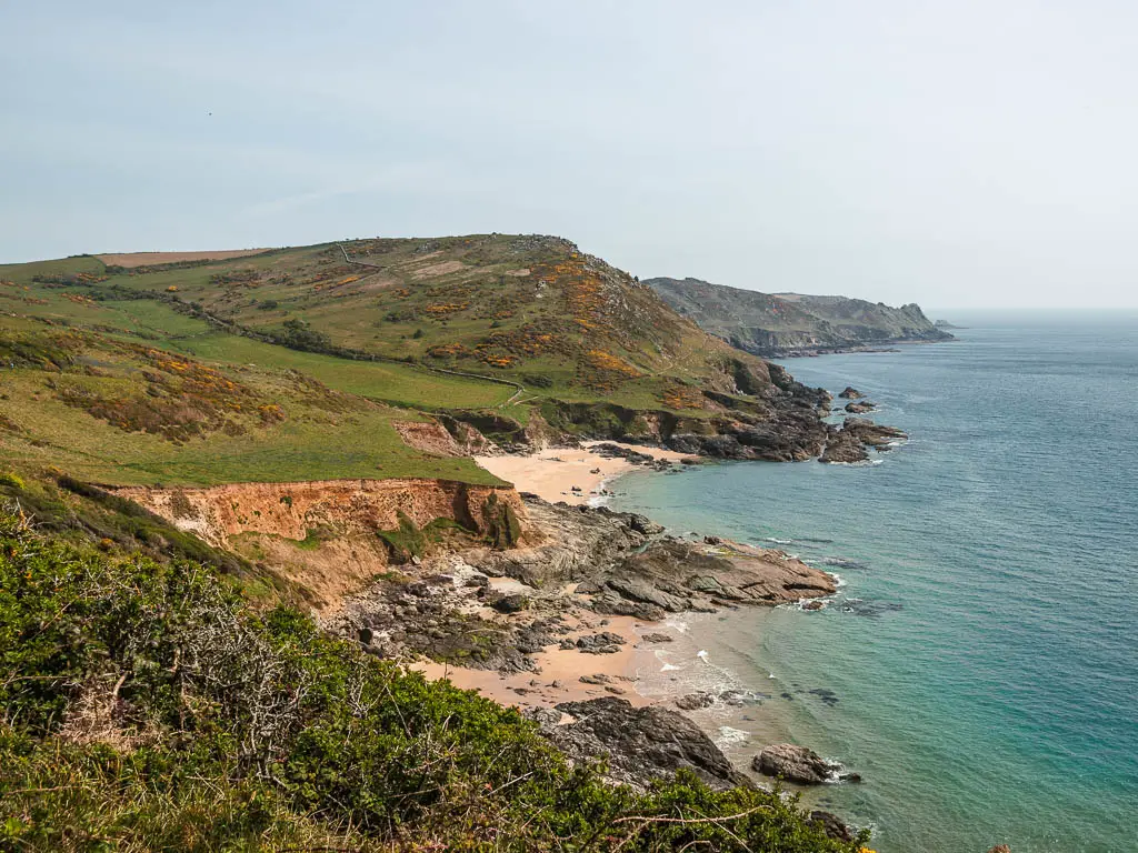 Looking along the rugged cliff coastline on the walk from Salcombe to Gara Rock. The cliff top on the left is made up of undulating grass hills. At the bottom of the cliffs are a couple of sandy beach coves. The sea is blue and becomes turquoise as it meets the sand.