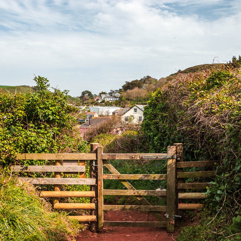 A wooden fence and gate, surrounded by bushes, with a few houses visible further ahead.