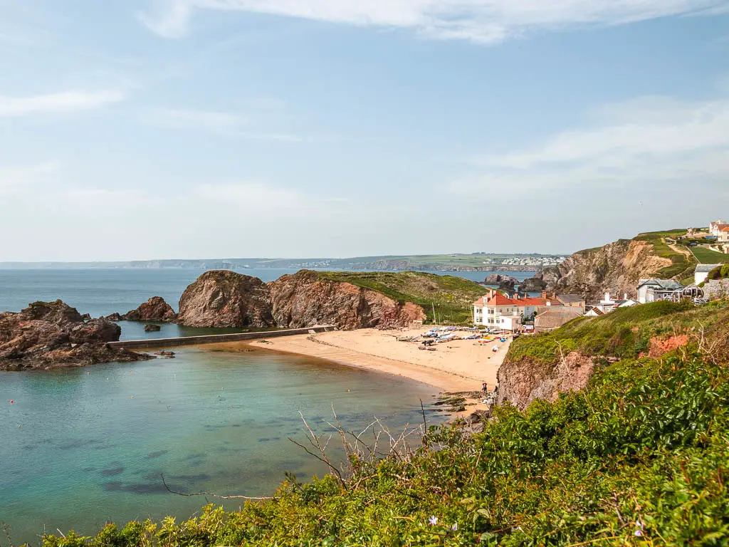Looking over the hedge to Hope cove at the end of the walk from Salcombe. The cove is made up of a small sandy beach on the right, with rocks around the side and into the blue turquoise sea. There is a cluster of houses behind the beach.
