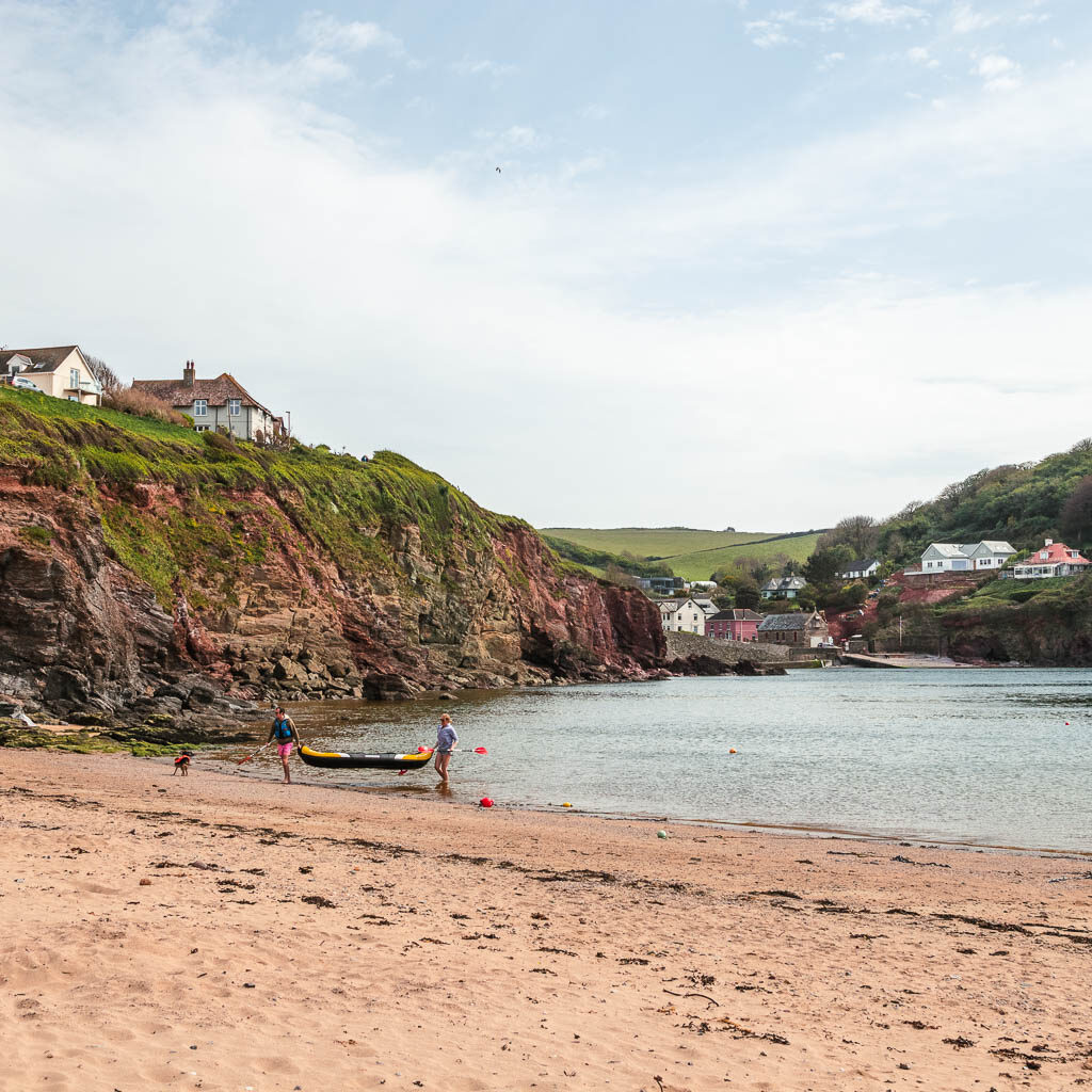 A sandy beach with cliffs to the left and the sea ahead. There are a couple of people carrying a kayak out of the sea.