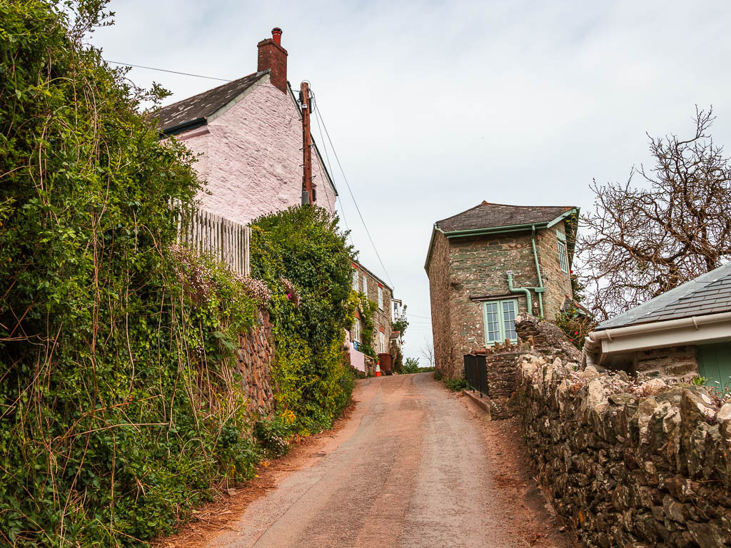 A country road leading uphill, with a stone wall on the right, and bushes covering the stone wall on the left. there are some houses ahead at the top of the road. 