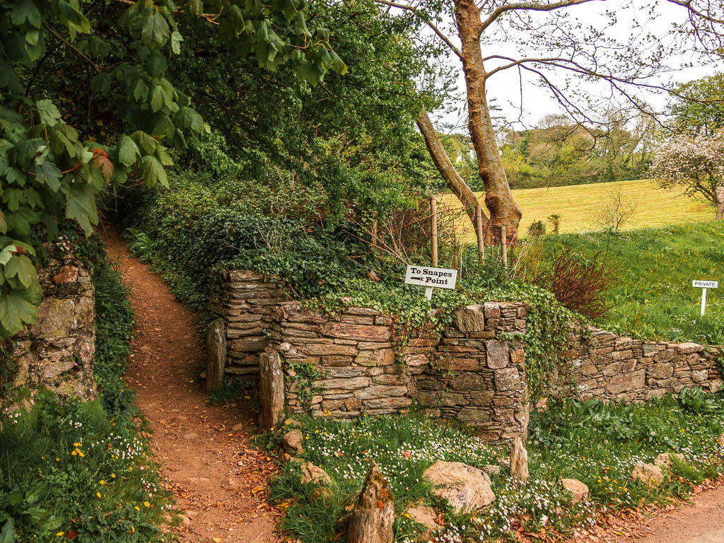 A dirt trail leading up to the left through a stone wall with a sign pointing to Snapes Point, on the walk route from Salcombe. 