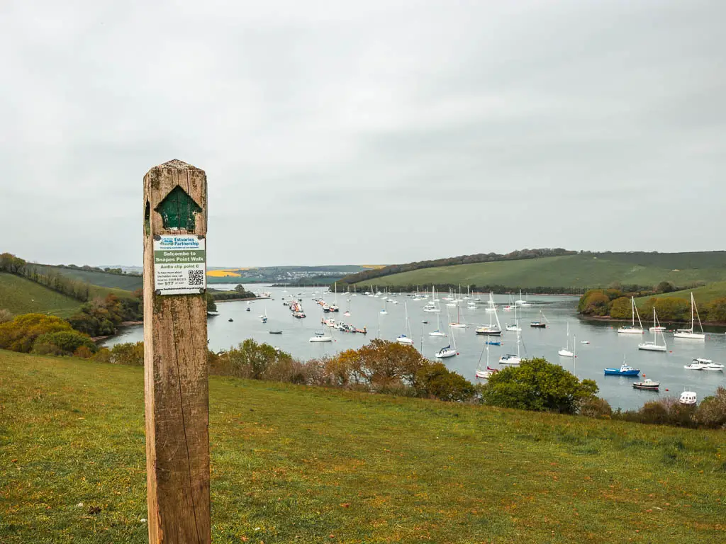 A wooden trail signpost on the green hill field, with a green arrow pointing ahead. The estuary with lots of boats is visible ahead. 