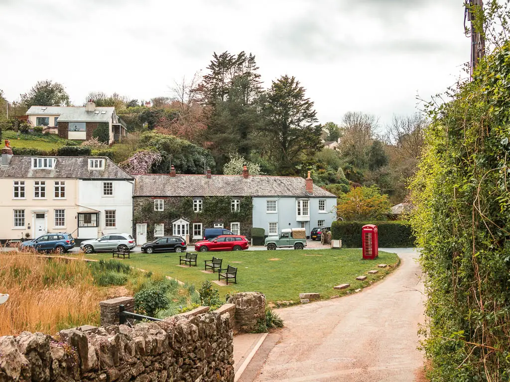 Looking down the road to a green with some houses on the other side at the end of the circular walk from Salcombe to Snapes Point. There are a few wooden benches on the green and a red telephone box. There are cars parked along the edge of the green.