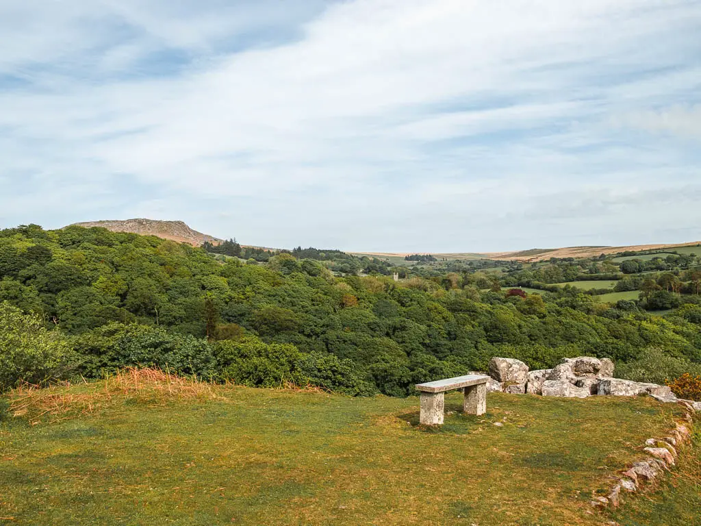 A stone bench on a flat grass hill top at the end of the Burrator Reservoir circular walk. There is a view across the tree tops to Sheepstor looming above in the distance.