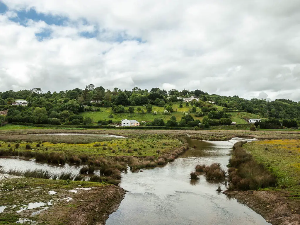 Looking along the creek and boggy ground towards a hill in the distance, the the Casltock Riverside walk. There are trees dotted about the hill, and a house at the bottom of it.