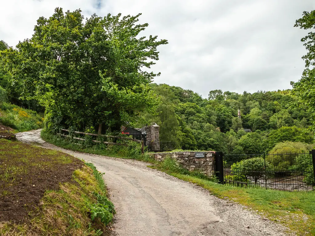 A country road curving uphill to the left then right. There is a stone wall and wooden fence on the other side of the road, and lots of bushy trees tops visible beyond that,.