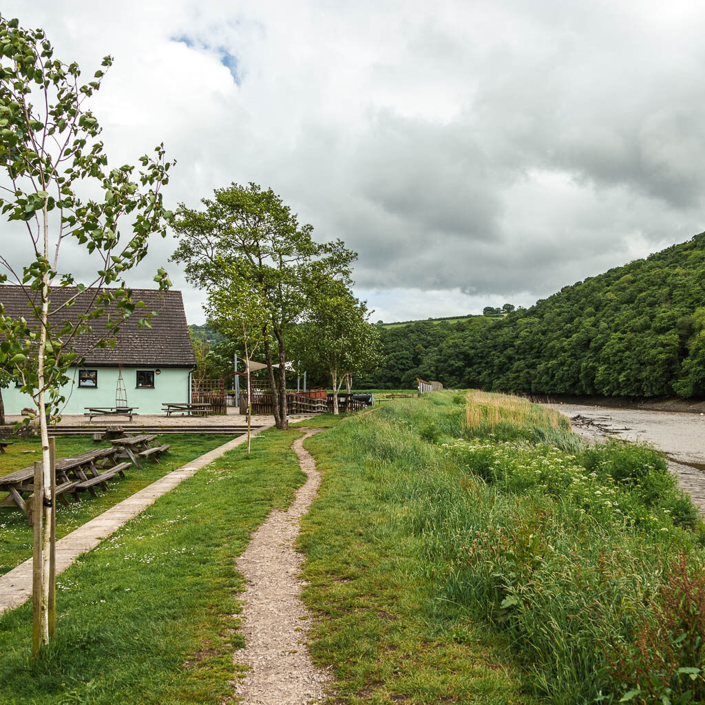 A narrow trail running along the grass next to the river. There is a castle light blue building ahead to the left of the trail.