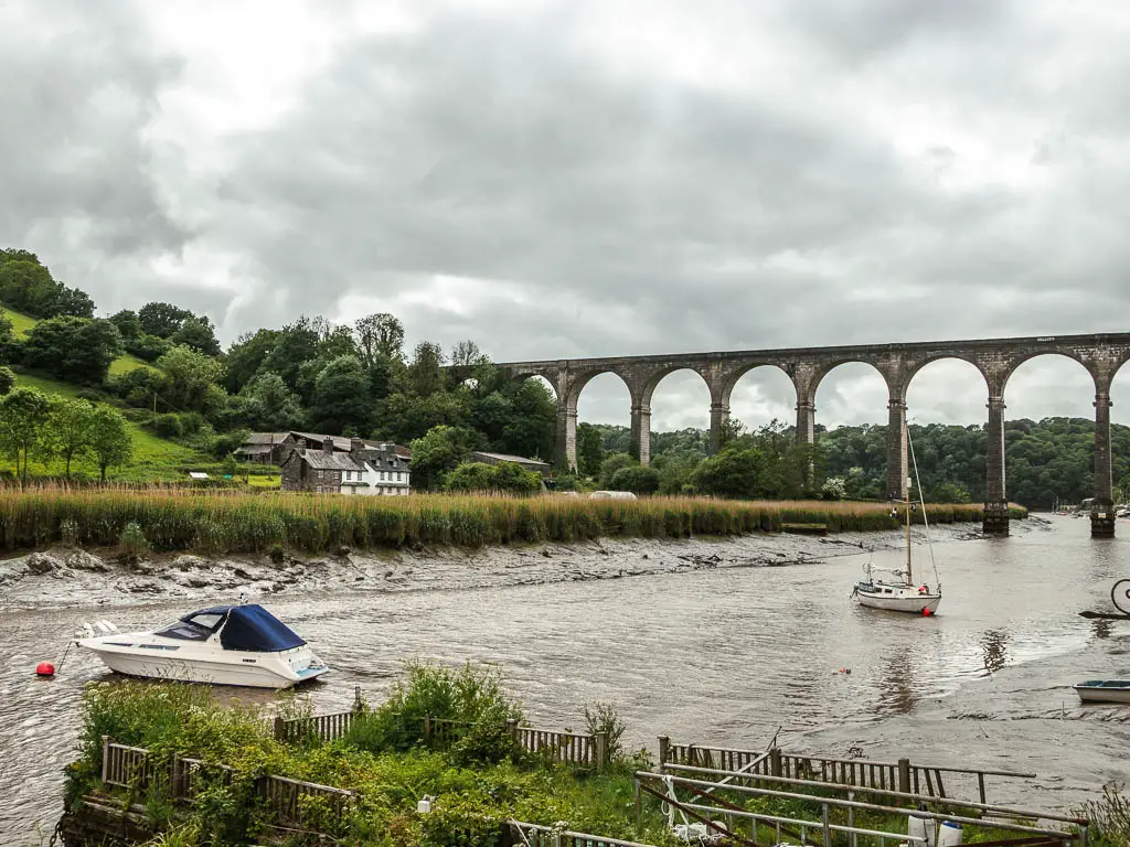 Looking down along the river Tamer towards the viaduct at the start of the riverside walk. There tide is low and there are a couple of small boats on the river. There is a field and hill on the other side, and a house next to one of the arches of the viaduct. 