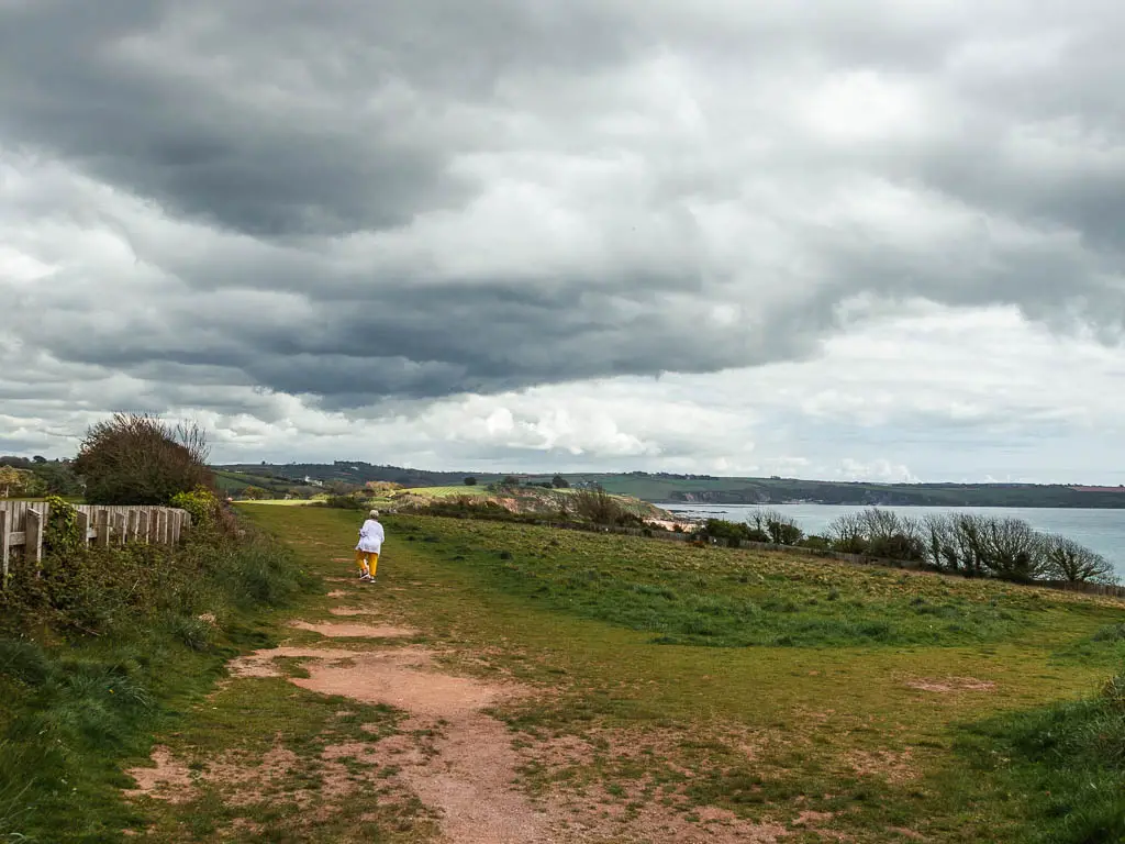 A large green with a dirt path on the left and a person walking along it. There is a bit of a sea view to the right.