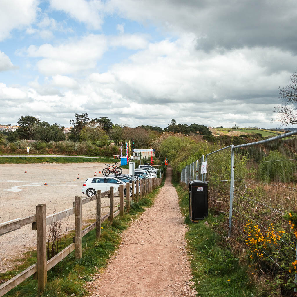 A path running down the left side of a car park. There are a few cars parked ahead. 