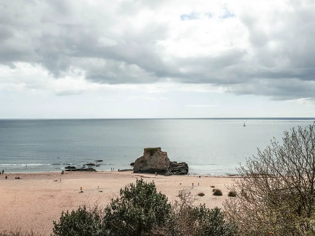 Looking down to the sandy beach as it meets the sea. There is a large rock on the sand. There are a few people walking on the beach.