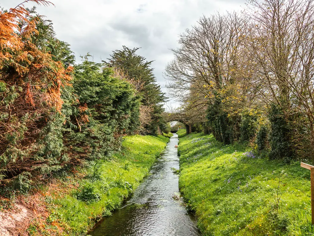 A long stream of water, with grass banks, and the arch of a stone bridge just visible ahead. There are trees to the left and a stone wall and less trees to the right.