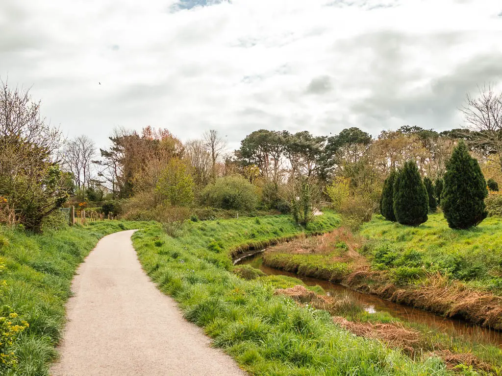 A wide neat path on the left and a stream to the right. The path is lined with grass and there are trees up ahead. 