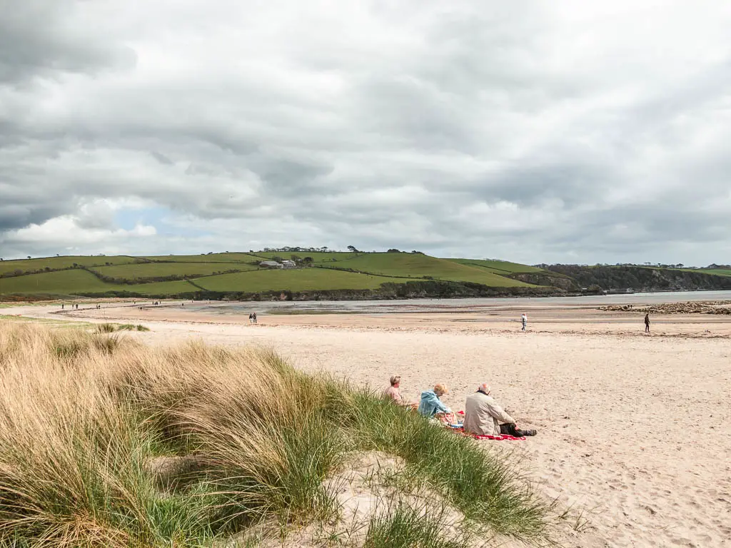 Par Sands Beach at the end of the walk from Charlestown. The beach is long with light coloured sand, and a view to the hills on top of the cliff on the other side. There are a few people sitting on the beach in front of some small sand dunes. 