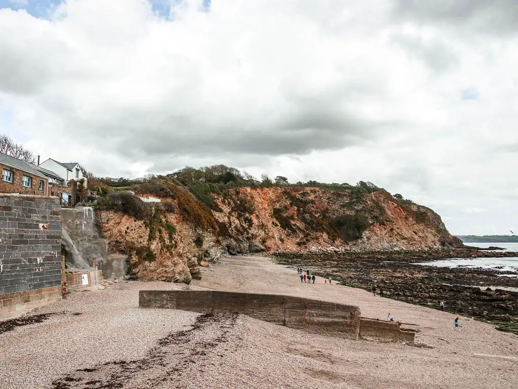 Looking down the sandy beach in Charlestown, at the start of the coastal walk to Par. There are rugged cliffs on the other side of the beach. There are a few people walking on the beach.