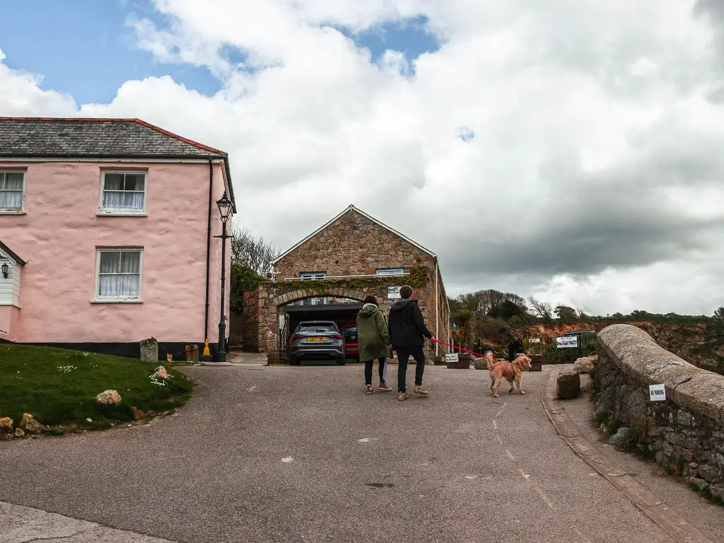 A road going uphill, with a light pink house on the left, a stone wall on the right, and a couple of people walking their dog up the road. 