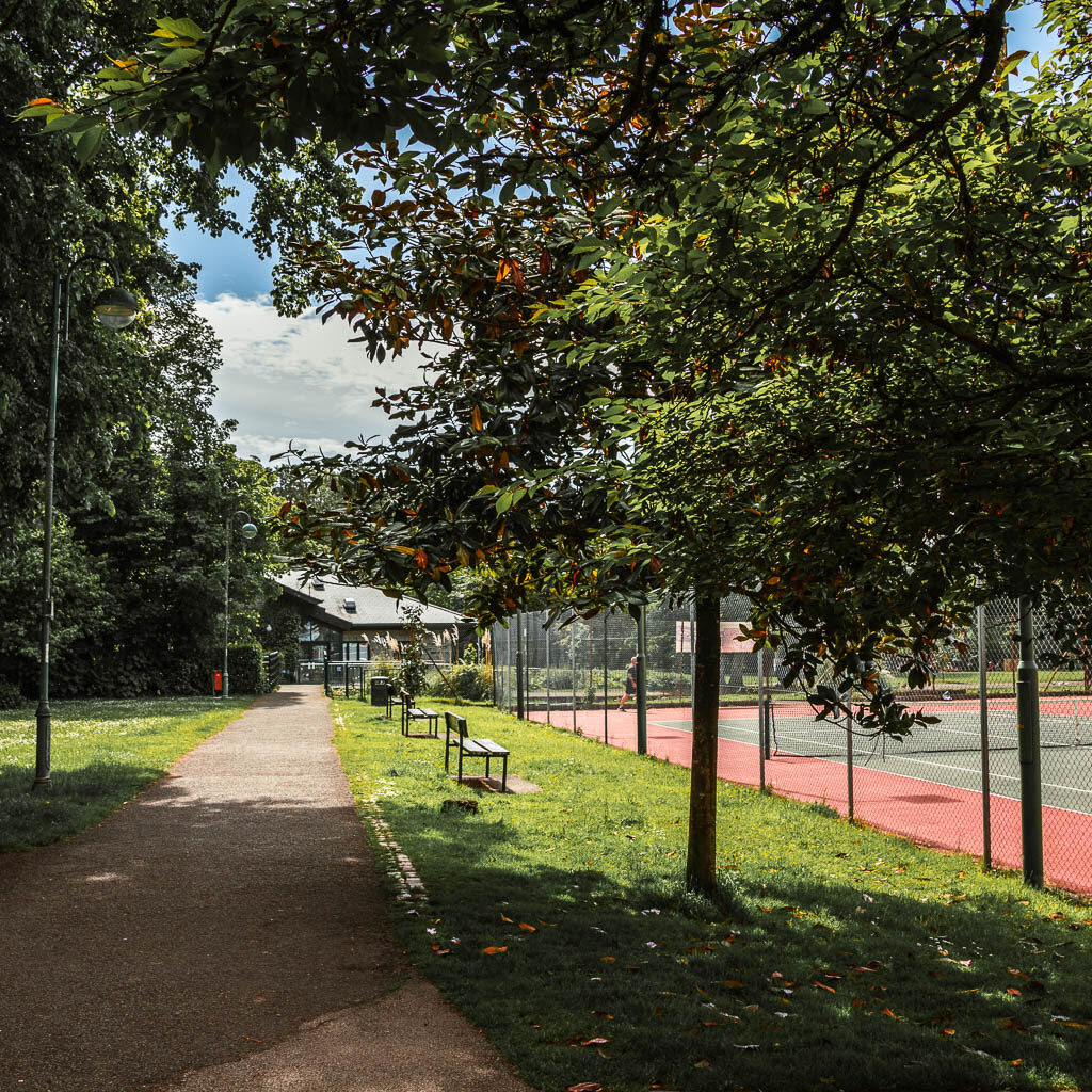 a path on the left and a patch of green on the right leading to some tennis courts. There are woodmen benches on the green.