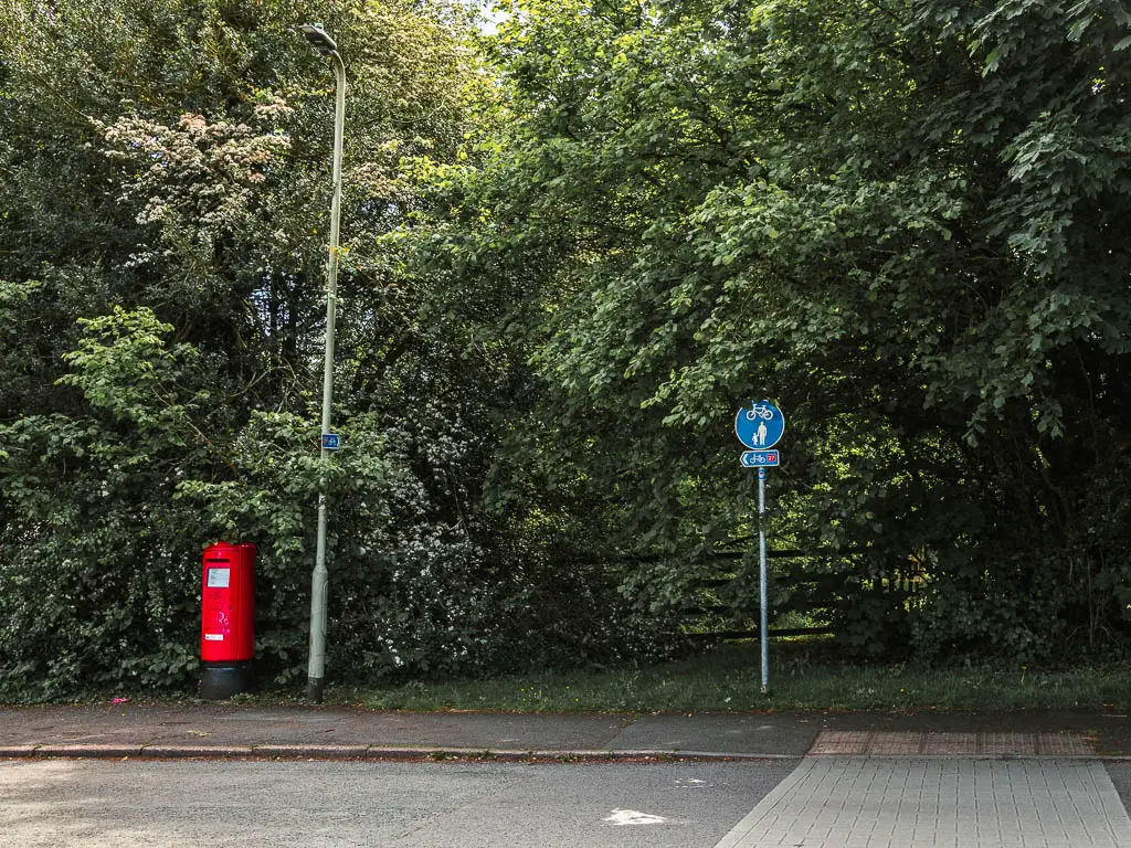 Looking across the road to a red post box and a blue walking and cycling signpost, infant of the bushes and trees.