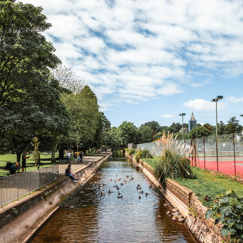 Looking along the river, with a path on the left and tennis courts on the right. There are ducks on the river. 