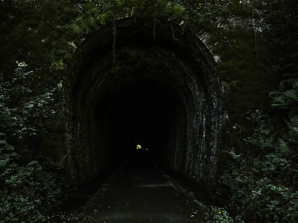 Looking through a dark and long stone walled tunnel with a light in the distance at the end of it, on the Drakes trail walk. 