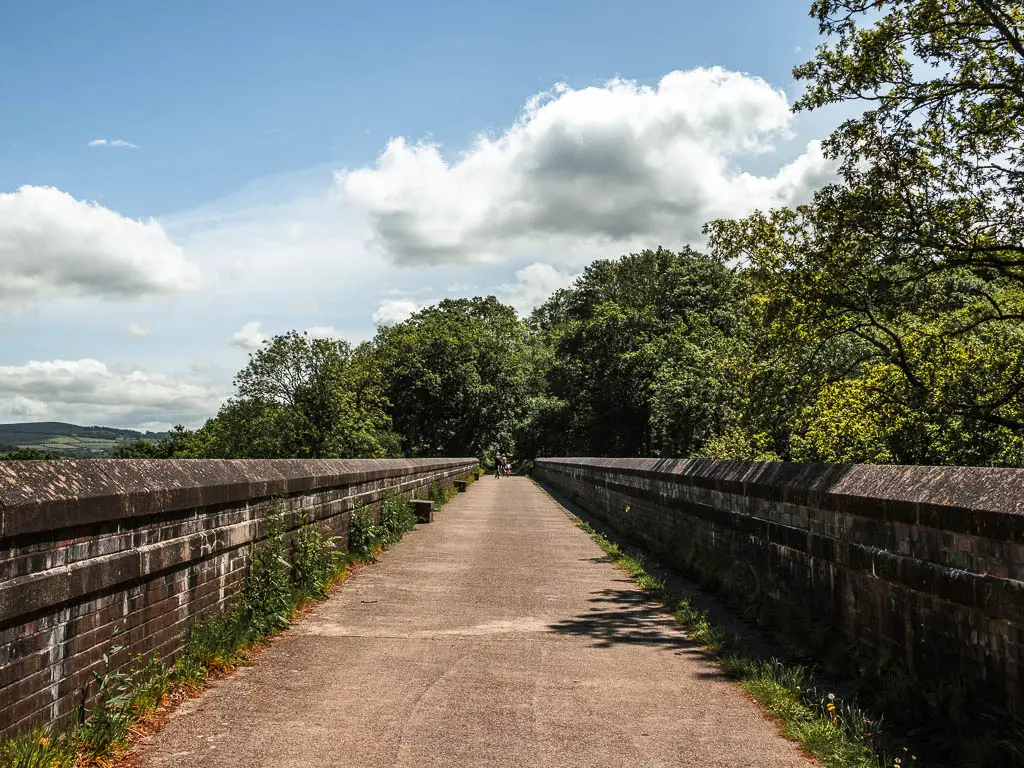 Looking along a wide bridge path with  stones walls on both side. The path leads to woodland on the other side.