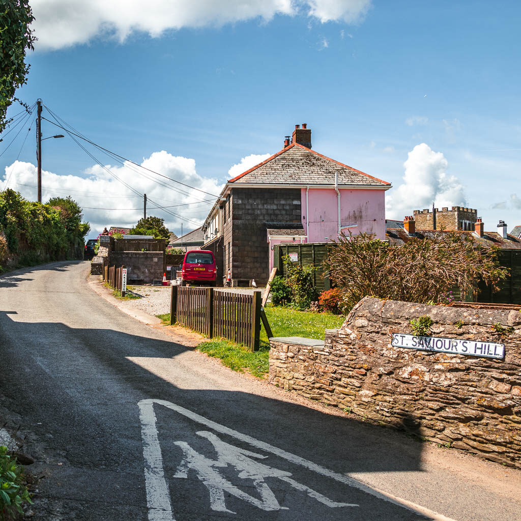 A road with a tone wall and road sign saying St Savours Hill. There is a pink coloured house further along the side of the road.