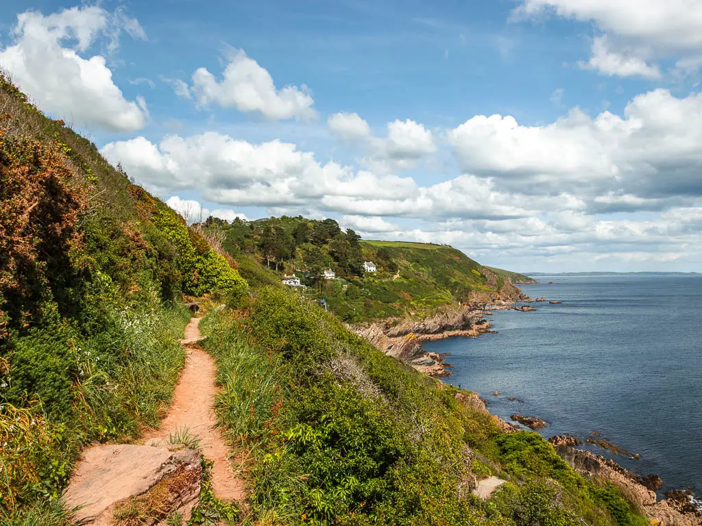 A view across towards Polperro, at the end of the walk from Fowey. There is a dirt trail on the left, on the side of a grass covered hill, with the blue sea on the right and a view to a few white houses on the cliff edge ahead.