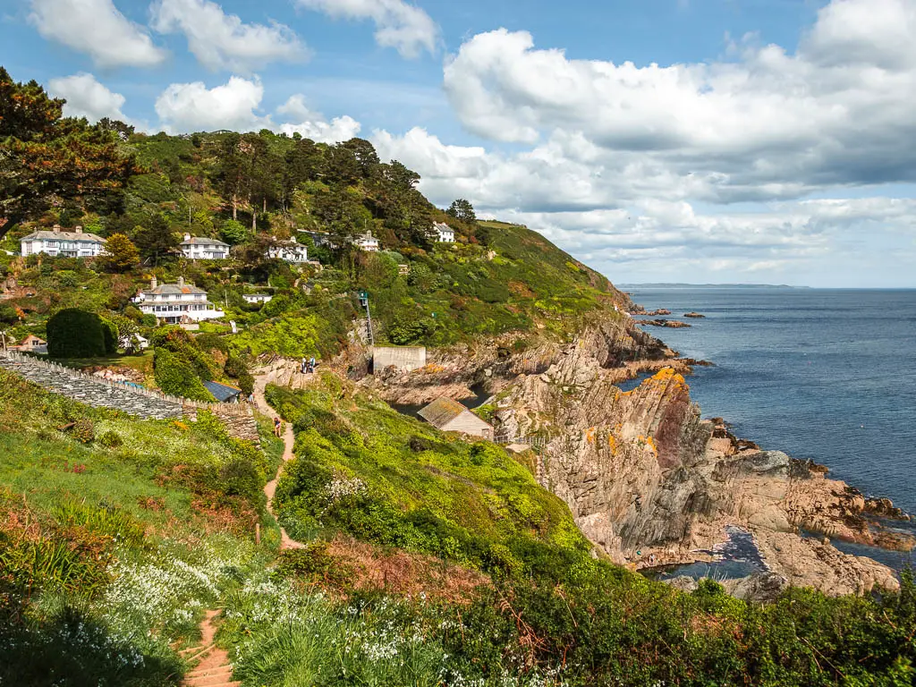 Looking along the pretty grass covered cliff, towards Polperro, with a view to some jagged pointy rocks ahead on the right and some white houses on the hill on the other side at the end of the coastal walk from Fowey.