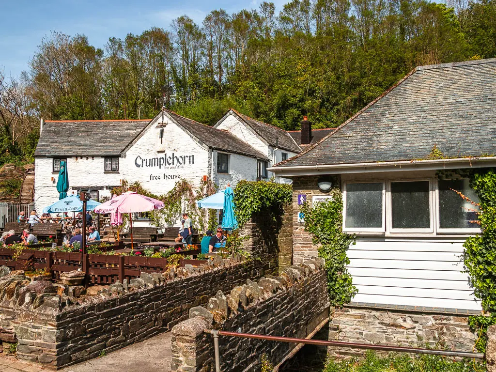 The white walled Crumplehorn Pub ahead to the left and on the right, the public toilets with a bridge and stone wall leading to it.