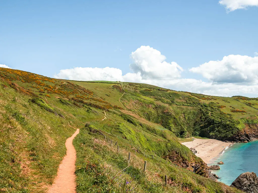 A narrow trail on the left, partway up a grass hill along the coastal walk from Fowey to Polperro. The hill leads down to the right and to a small sandy beach cove.