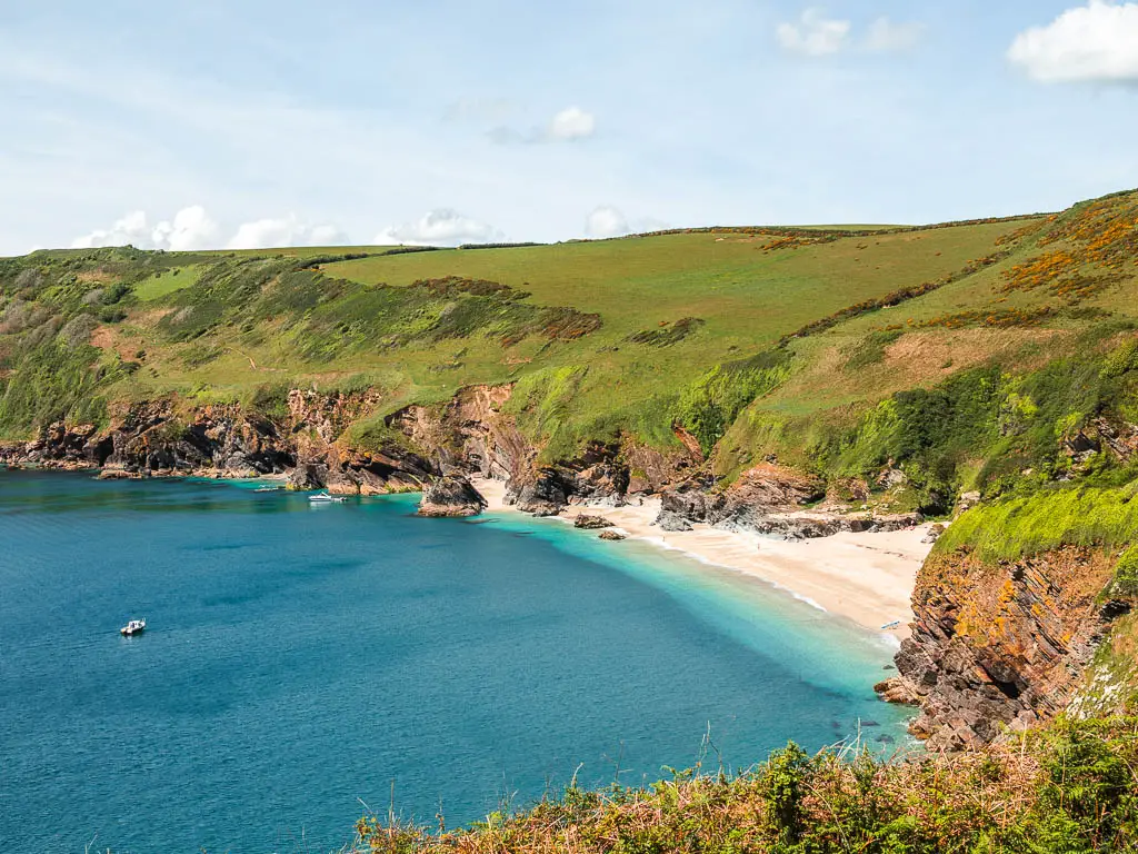 Looking down to a secluded beach cove on the right on the coastal Fowey to Polperro walk. The beach is surrounded by cliffs with a grass covered top. The sea is light blue and turquoise as it meets the beach. There is a small boat in the water.