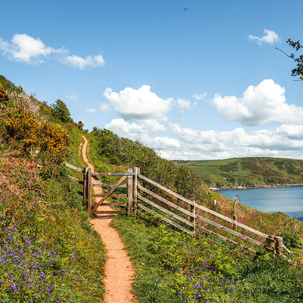 A trail leading through a wooden gate and fence on the hill side, with a small view to the sea to the right.