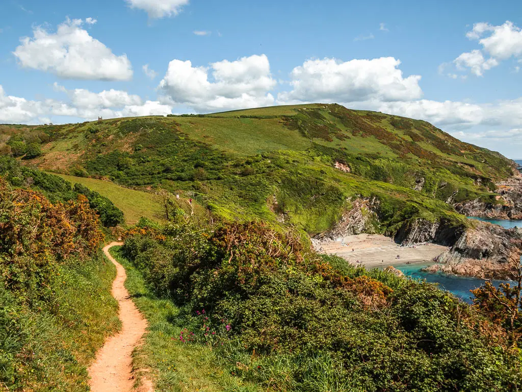 A trail on the left leading along the top of the grass and bush covered cliff on the Fowey to Polperro coastal walk. There is a view to a small sandy beach cove down to the right.