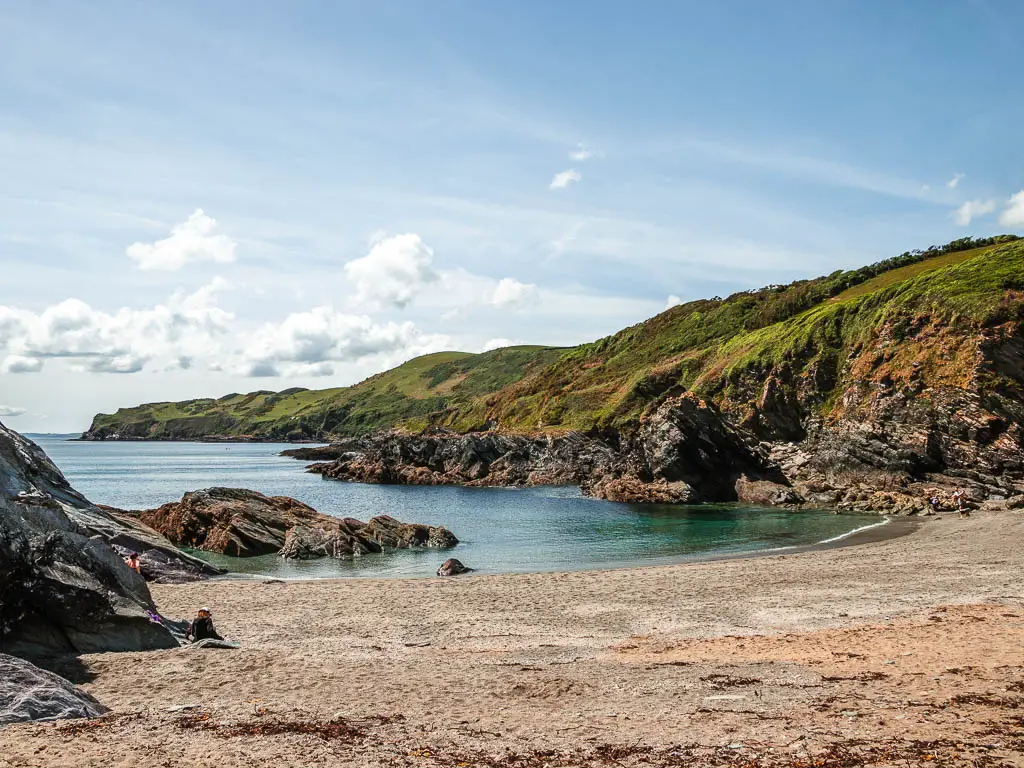 Looking across the sand beach cove, towards the sea and rocky cliffs with a grass covered clifftop on the other side.