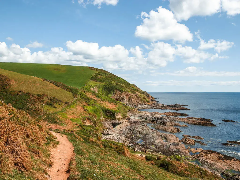 A dirt trail on the left side, running along the clifftop along the coastal Fowey to Polperro walk. The trail continues along the undulaing grass covered clifftop, and then the cliff forms rocks where it meets the sea below.