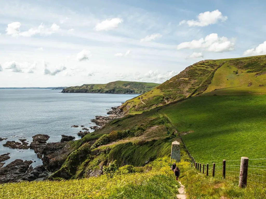Looking down hill along the trail, and then along the undulating clifftop on the Fowey to Polperro Coastal walk. Where the cliff meets the sea on the left it forms lots of black rocks. There are a couple of people walking up the trail.