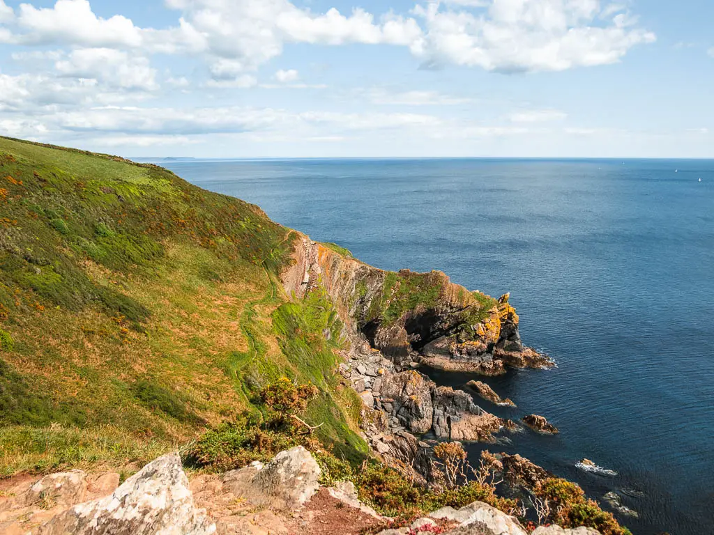 Looking down the grass covered cliff side to rocky, jagged cliff outcrops at the bottom where the cliff meets the blue sea.