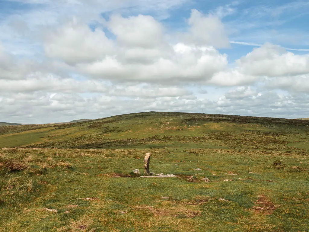 Looking across the grass to the hill in the distance on the Ivybridge to Western Beacon circular walk. There is a thin rock standing up on the grass. 